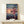 May include: Framed Spirited Away movie poster showing animated girl with brown hair in pink outfit running across flooded train tracks at sunset, displayed on light wood shelf next to small white wrapped gift with greenery, against cream wall in minimalist room setting