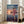 May include: Framed Studio Ghibli Spirited Away movie poster leaning against white wall in bright living room. Shows anime girl with brown hair in pink outfit running on wooden railway tracks over water at sunset. Japanese and English text visible. Beige sofa and windows with natural light in background.