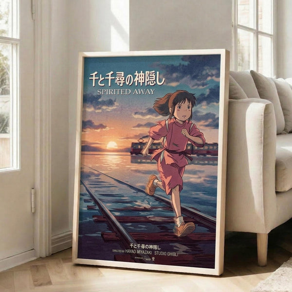 May include: Framed Studio Ghibli Spirited Away movie poster leaning against white wall in bright living room. Shows anime girl with brown hair in pink outfit running on wooden railway tracks over water at sunset. Japanese and English text visible. Beige sofa and windows with natural light in background.