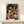 May include: Framed Spirited Away movie poster displayed on light wood shelf next to small white wrapped gift. Poster features young girl in red outfit surrounded by colorful animated characters in traditional Japanese street scene with paper lanterns. Artwork has vibrant colors with Japanese and English text at top. Natural lighting on white wall background.