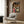 May include: Framed Spirited Away movie poster mounted on wood-paneled wall above modern beige armchair with geometric pillow. Poster shows young girl in pink kimono surrounded by animated characters including No-Face and colorful spirits. Contemporary living room setting with brass floor lamp, dark round side table with open book, and neutral tones throughout.