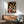 May include: Framed Spirited Away movie poster hanging on white wall above wooden sideboard with table lamp. Poster shows young girl in red outfit with various animated characters in traditional Japanese street scene with lanterns. Mid-century modern furniture including teal upholstered chair with wooden arms and round coffee table visible in foreground. Natural sunlight creates geometric shadows across the minimalist room setting.