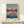 May include: Framed Spirited Away anime poster displayed on light wood shelf with small wrapped gift. Poster shows young girl in pink dress standing on flooded railway tracks at sunset, with Japanese and English text. Natural wood frame against cream wall in minimalist styling.