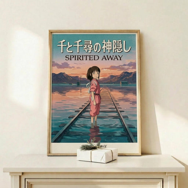 May include: Framed Spirited Away anime poster displayed on light wood shelf with small wrapped gift. Poster shows young girl in pink dress standing on flooded railway tracks at sunset, with Japanese and English text. Natural wood frame against cream wall in minimalist styling.