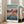 May include: Framed poster of animated character in red dress standing on flooded railway tracks at sunset, with Japanese and English text reading "Spirited Away." Poster leans against white wall next to beige upholstered chair in bright living room with large windows. Art features turquoise water, purple-orange sky, and mountains in background.