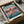 May include: Framed Spirited Away movie poster showing young girl in pink dress walking on flooded railway tracks at sunset, with Japanese and English text at top. Poster displayed in black frame inside cardboard shipping box with protective packaging materials on wooden surface.