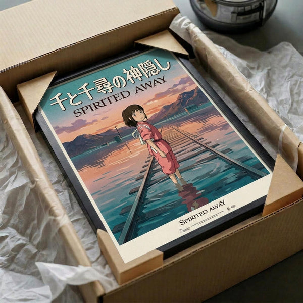 May include: Framed Spirited Away movie poster showing young girl in pink dress walking on flooded railway tracks at sunset, with Japanese and English text at top. Poster displayed in black frame inside cardboard shipping box with protective packaging materials on wooden surface.