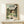 May include: Framed Spirited Away movie poster in light wood frame displayed on white wooden shelf against cream wall. Poster shows anime girl with brown bob haircut in green and white striped shirt standing in traditional Japanese garden setting with stone lanterns, trees, pond, and wooden buildings. Small white decorative box sits on shelf in front of frame.