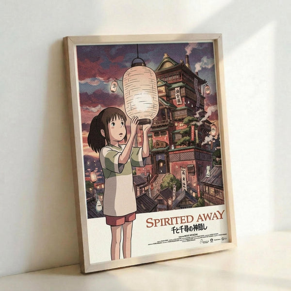 May include: Framed Spirited Away movie poster leaning against white wall. Features anime character Chihiro holding white paper lantern with fantastical multi-story bathhouse building behind her against purple twilight sky. Artwork shows Studio Ghibli animation style with detailed architectural elements and warm lighting. Light wooden frame, appears medium-sized print in portrait orientation.