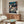 May include: Framed Spirited Away movie poster hanging above wooden credenza in modern living room. Shows young girl with dark hair surrounded by white spirits and traditional Japanese architecture. Poster features vibrant blues and warm lighting. Room has cream walls, white upholstered chair, glass coffee table, and minimalist decor with natural wood finishes.