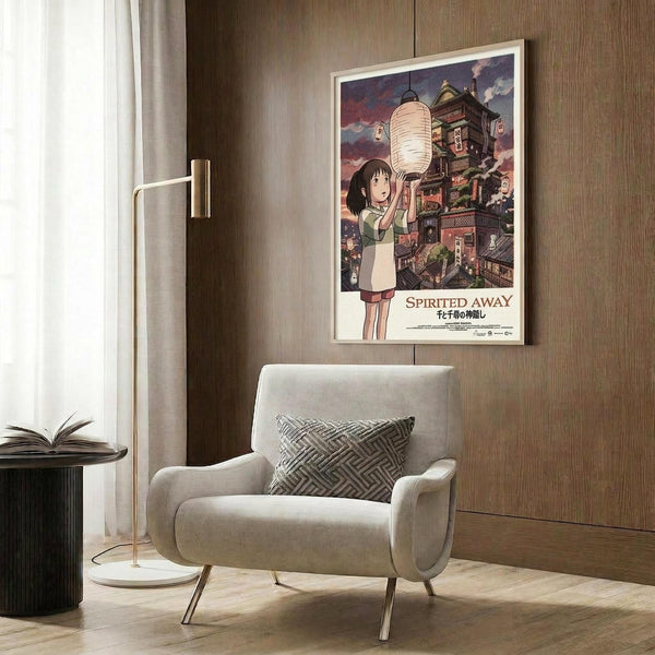 May include: Framed Spirited Away movie poster mounted on wood-paneled wall above beige armchair with geometric pillow. Poster shows anime girl with paper lantern against traditional Japanese building backdrop with purple sky. Modern reading corner with gold floor lamp and open book on dark round table.