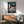 May include: Framed Spirited Away movie poster hanging on white wall above wooden sideboard with table lamp. Features anime girl with dark hair surrounded by white spirits and traditional Japanese bathhouse architecture in background. Mid-century modern living room setting with teal upholstered chair and round coffee table. Natural sunlight creates geometric shadows on wall. Poster shows vibrant blues and earth tones in detailed illustration style.