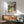 May include: Framed Studio Ghibli Spirited Away movie poster featuring anime character Chihiro in green and white shirt standing in traditional Japanese garden setting with stone lanterns and pathways. Poster displayed above mid-century modern wooden credenza with table lamp, next to sage green upholstered armchair with wooden frame in bright, naturally-lit room with white walls.