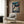 May include: Framed Spirited Away movie poster hanging on wood-paneled wall above beige upholstered armchair with geometric patterned throw pillow. Poster shows young girl character surrounded by white spirits and traditional Japanese architecture in rich blues and warm tones. Modern floor lamp with brass finish and circular base stands nearby next to small round table with open book.