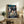 May include: Framed Studio Ghibli Spirited Away movie poster displayed on rustic wooden sideboard in cozy living room setting. Poster features young girl character surrounded by white spirits and traditional Japanese architecture with red lanterns. Natural wood frame, positioned next to fern plant, ceramic vase, and stacked books. Beige armchair visible in foreground.