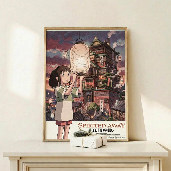 May include: Framed Spirited Away movie poster featuring Chihiro holding a white paper lantern with the ornate bathhouse building behind her against a purple twilight sky. The wooden-framed print sits on a light wooden shelf next to a small white wrapped gift with greenery. The anime artwork shows rich colors and Studio Ghibli's distinctive illustration style with Japanese text below the English title.