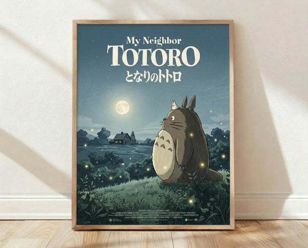 May include: Framed poster print leaning against white wall on wooden floor. Features Totoro character in moonlit nighttime scene with small houses in background, surrounded by glowing fireflies. Blue-green gradient sky with full moon, lush vegetation in foreground. Japanese text below English title. Natural wood frame, soft window light casting shadows on wall.