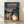 May include: Framed poster print showing iconic Studio Ghibli characters Totoro and a young girl with umbrellas standing at a rural bus stop. The artwork features a twilight scene with power lines, trees, and a distant bus, rendered in animated film style with muted blues, greens, and warm sunset tones. The wooden frame sits on light wood flooring against a white wall with natural lighting creating soft shadows.
