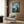 May include: Studio Ghibli-inspired poster featuring Totoro character in nighttime forest scene with full moon and fireflies, displayed in light wooden frame on warm wood paneled wall above beige upholstered armchair with geometric patterned throw pillow, modern floor lamp, and dark round side table with open book