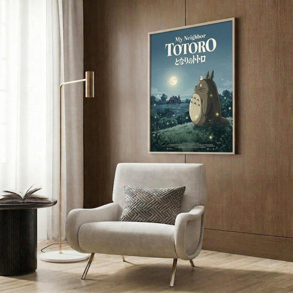 May include: Studio Ghibli-inspired poster featuring Totoro character in nighttime forest scene with full moon and fireflies, displayed in light wooden frame on warm wood paneled wall above beige upholstered armchair with geometric patterned throw pillow, modern floor lamp, and dark round side table with open book