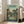 May include: Large framed poster leaning against white wall next to beige sofa in bright living room. Poster shows Japanese village scene with traditional houses, power lines, large tree, and three small figures including Totoro character walking on dirt path. Green and yellow tones with Japanese text at top, English title "My Neighbor Totoro" and Studio Ghibli credit below. Natural wood frame, approximately 2-3 feet tall.