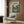 May include: Vintage-style My Neighbor Totoro movie poster mounted on wood-paneled wall above beige upholstered armchair. Poster features Japanese village scene with traditional houses, power lines, trees, and Totoro characters in muted green and brown tones. Modern reading nook setting with geometric pillow, brass floor lamp, and dark side table with open book.
