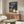 May include: Studio Ghibli My Neighbor Totoro poster displayed above a mid-century modern wooden credenza in a minimalist living room. The poster shows the iconic gray Totoro character with an umbrella standing next to a young girl at a rural bus stop, surrounded by lush green trees against a twilight sky. Features Japanese text at the top. Room includes a white curved chair, oval glass coffee table, and modern lamp with warm wood tones throughout.