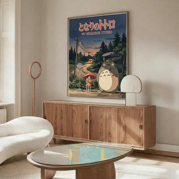 May include: Studio Ghibli My Neighbor Totoro poster displayed above a mid-century modern wooden credenza in a minimalist living room. The poster shows the iconic gray Totoro character with an umbrella standing next to a young girl at a rural bus stop, surrounded by lush green trees against a twilight sky. Features Japanese text at the top. Room includes a white curved chair, oval glass coffee table, and modern lamp with warm wood tones throughout.