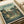 My Neighbor Totoro vintage-style poster print featuring Japanese characters and English text at top, illustrated scene of traditional Japanese neighborhood with wooden houses, power lines, and trees in muted green and brown tones. Two small children with umbrella stand on a path next to large white Totoro character. Studio Ghibli and Hayao Miyazaki credits appear at bottom. Print shown flat on white surface with rolled paper tube nearby.