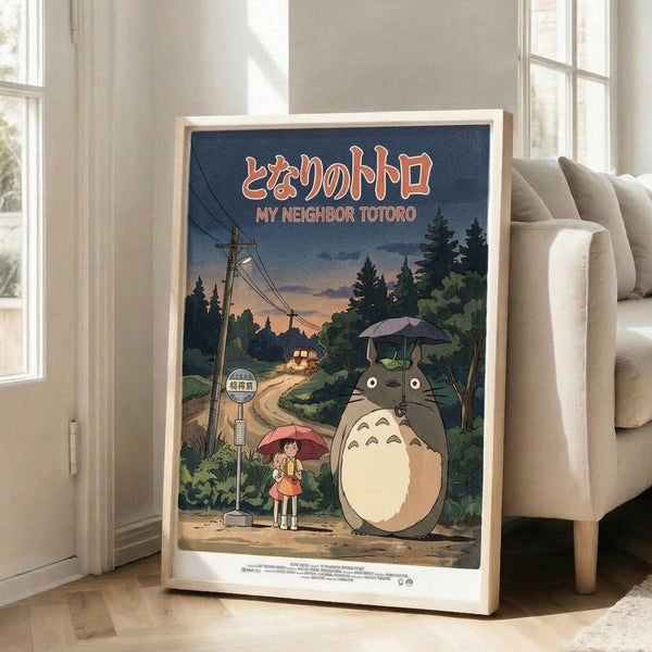 May include: Framed poster displayed in bright living room next to beige sofa. Artwork shows iconic scene from Japanese animated film with large gray forest creature holding umbrella standing beside young girl with red umbrella at rural bus stop. Features power lines, trees, dirt road, and Japanese text at top. Vintage movie poster style with rich blues, greens, and warm sunset colors in natural wood frame on hardwood floor.