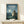 May include: Framed poster featuring Totoro character standing in moonlit countryside with glowing fireflies, houses in background, and Japanese text. Blue-green evening sky with full moon. Natural wood frame displayed on light wooden shelf beside small wrapped white gift box against cream wall.