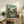 May include: Framed Studio Ghibli My Neighbor Totoro movie poster displayed on rustic wooden sideboard in cozy living room setting. Poster features Japanese village scene with traditional houses, power lines, large tree, and small figures including Totoro character in muted green and beige tones. Scene includes fern plant, ceramic vase, books, and brown leather furniture creating warm, nostalgic atmosphere with natural lighting.