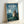 May include: Framed poster print in light wood frame leaning against white wall. Features Totoro character standing in moonlit countryside scene with glowing fireflies, houses in distance, and full moon in blue-green night sky. Title text "My Neighbor TOTORO" in white letters with Japanese characters below. Soft lighting creates gentle shadows on wall.