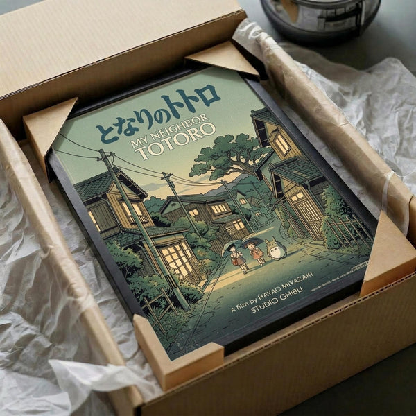 May include: Framed My Neighbor Totoro poster in wooden frame, nestled in cardboard shipping box with protective white tissue paper. The artwork shows a Japanese neighborhood scene with traditional houses, power lines, trees, and small figures including Totoro characters in muted green and brown tones. Frame appears medium-sized relative to the packaging materials surrounding it.