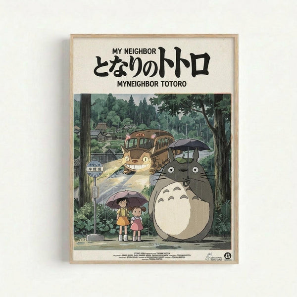 Framed poster featuring Totoro, a large gray forest spirit with white belly, standing under an umbrella in the rain alongside two young girls at a bus stop. The Catbus, a grinning cat-shaped vehicle, appears in the background among lush green trees and rural Japanese buildings. Japanese text appears at the top with English translation "My Neighbor Totoro." The artwork has Studio Ghibli's distinctive hand-drawn animation style with rich greens and earth tones, displayed in a light wood frame.
