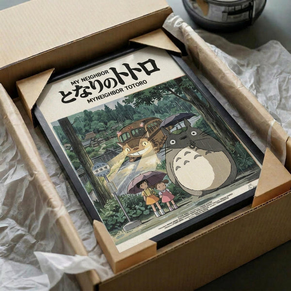 May include: Vintage-style poster featuring Totoro and Catbus characters from Studio Ghibli animation, displayed in black frame within cardboard shipping box with protective wrapping. Poster shows forest scene with children holding umbrellas beneath large grey Totoro and cat-shaped bus. Japanese text at top. Surrounded by white tissue paper and wooden protective strips.
