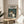 May include: Framed poster featuring Totoro and characters from the animated film, displayed on a round wooden side table next to a beige upholstered chair. The poster shows forest creatures including the iconic gray Totoro with white belly, smaller creatures, and children with umbrellas in a lush green woodland setting. Natural lighting creates soft shadows across the neutral-toned room with white walls and plant leaves visible in the corner.