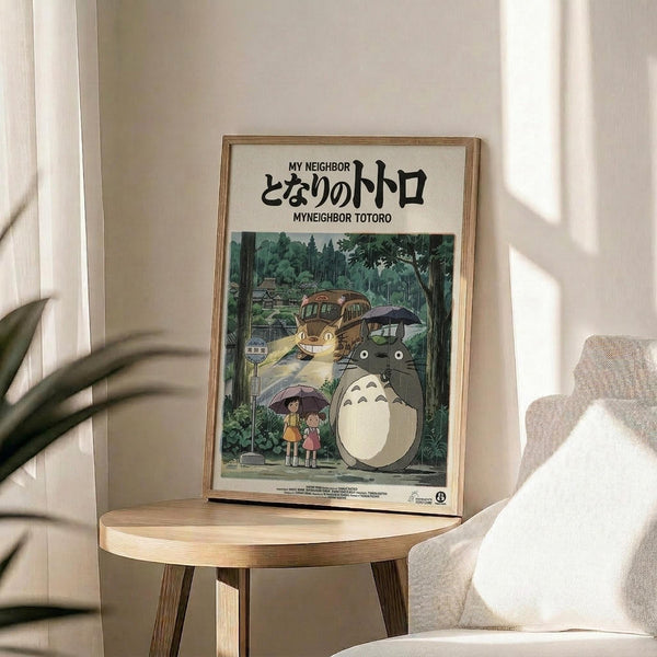 May include: Framed poster featuring Totoro and characters from the animated film, displayed on a round wooden side table next to a beige upholstered chair. The poster shows forest creatures including the iconic gray Totoro with white belly, smaller creatures, and children with umbrellas in a lush green woodland setting. Natural lighting creates soft shadows across the neutral-toned room with white walls and plant leaves visible in the corner.