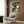 May include: A framed Studio Ghibli movie poster featuring Totoro, a large gray forest creature with an umbrella, standing beside two young girls under a massive green tree in a sunlit forest clearing. The poster hangs on a wood-paneled wall above a modern cream-colored armchair with geometric pillow, next to a brass floor lamp and dark round side table with an open book.