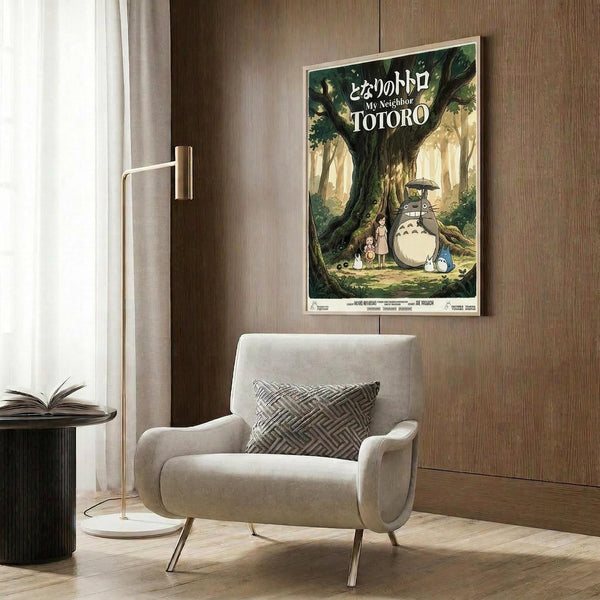 May include: A framed Studio Ghibli movie poster featuring Totoro, a large gray forest creature with an umbrella, standing beside two young girls under a massive green tree in a sunlit forest clearing. The poster hangs on a wood-paneled wall above a modern cream-colored armchair with geometric pillow, next to a brass floor lamp and dark round side table with an open book.