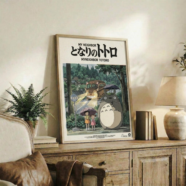 May include: Framed Studio Ghibli My Neighbor Totoro movie poster in light wood frame, displayed on rustic wooden sideboard with drawers. Scene shows iconic gray Totoro character with children under umbrellas at forest bus stop, plus Catbus. Styled with fern plant, cream lamp, ceramic vase, and books in cozy living room setting with beige armchair.