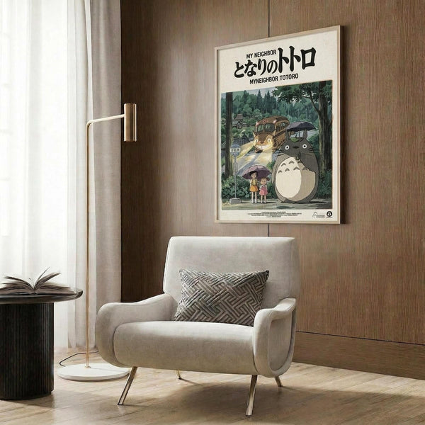 May include: A framed My Neighbor Totoro movie poster hanging on a wood-paneled wall above a beige upholstered armchair with geometric pillow. The vintage-style poster shows Totoro and characters under umbrellas in a forest scene with Japanese text. A brass floor lamp and dark side table with open book complete the cozy reading nook setup.