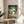 May include: Framed My Neighbor Totoro movie poster displayed on a round wooden side table next to a beige upholstered chair. The poster features the iconic gray Totoro character with children in a lush green forest setting. Natural lighting creates soft shadows across the minimalist room with white walls and a potted plant visible in the corner.