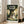 May include: Framed Studio Ghibli movie poster leaning against white wall next to beige sofa in bright room with windows. Poster shows Totoro character with umbrella standing beside two children under large forest tree, rendered in green and golden tones with Japanese text at top.