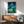 May include: Framed My Neighbor Totoro poster mounted on white wall above wooden credenza. Artwork shows nighttime scene with starry sky, crescent moon, children and large Totoro character in grassy field. Mid-century modern room setting with sage green armchair, round coffee table, and table lamp. Natural sunlight creates shadows across the space. Poster has thin brown frame.