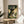 May include: Framed My Neighbor Totoro movie poster displayed on a round wooden side table next to a beige upholstered chair. The poster shows Totoro and children under a large tree in a sunlit forest scene with Japanese text at the top. Natural lighting creates soft shadows across the neutral-toned room setting with a plant visible in the corner.
