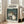 May include: Framed poster featuring beloved animated characters Totoro and Catbus in a lush forest setting with two children holding umbrellas at a bus stop. The artwork displays soft watercolor-style illustration in greens and earth tones, with Japanese text at top. Light wood frame leans against white wall next to beige sectional sofa in bright, naturally-lit modern living room with hardwood floors.