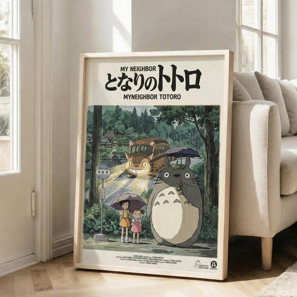 May include: Framed poster featuring beloved animated characters Totoro and Catbus in a lush forest setting with two children holding umbrellas at a bus stop. The artwork displays soft watercolor-style illustration in greens and earth tones, with Japanese text at top. Light wood frame leans against white wall next to beige sectional sofa in bright, naturally-lit modern living room with hardwood floors.