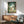 May include: Framed My Neighbor Totoro movie poster displayed above mid-century modern wooden credenza in living room setting. Features Totoro character with children in lush green forest scene. Sage green upholstered chair with wooden arms and small white table lamp visible. Warm natural lighting creates shadows on white wall. Wood-toned frame complements furniture's walnut finish.