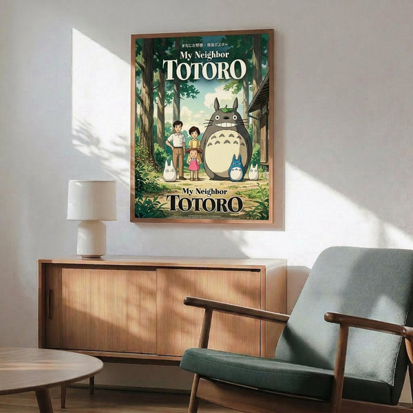May include: Framed My Neighbor Totoro movie poster displayed above mid-century modern wooden credenza in living room setting. Features Totoro character with children in lush green forest scene. Sage green upholstered chair with wooden arms and small white table lamp visible. Warm natural lighting creates shadows on white wall. Wood-toned frame complements furniture's walnut finish.